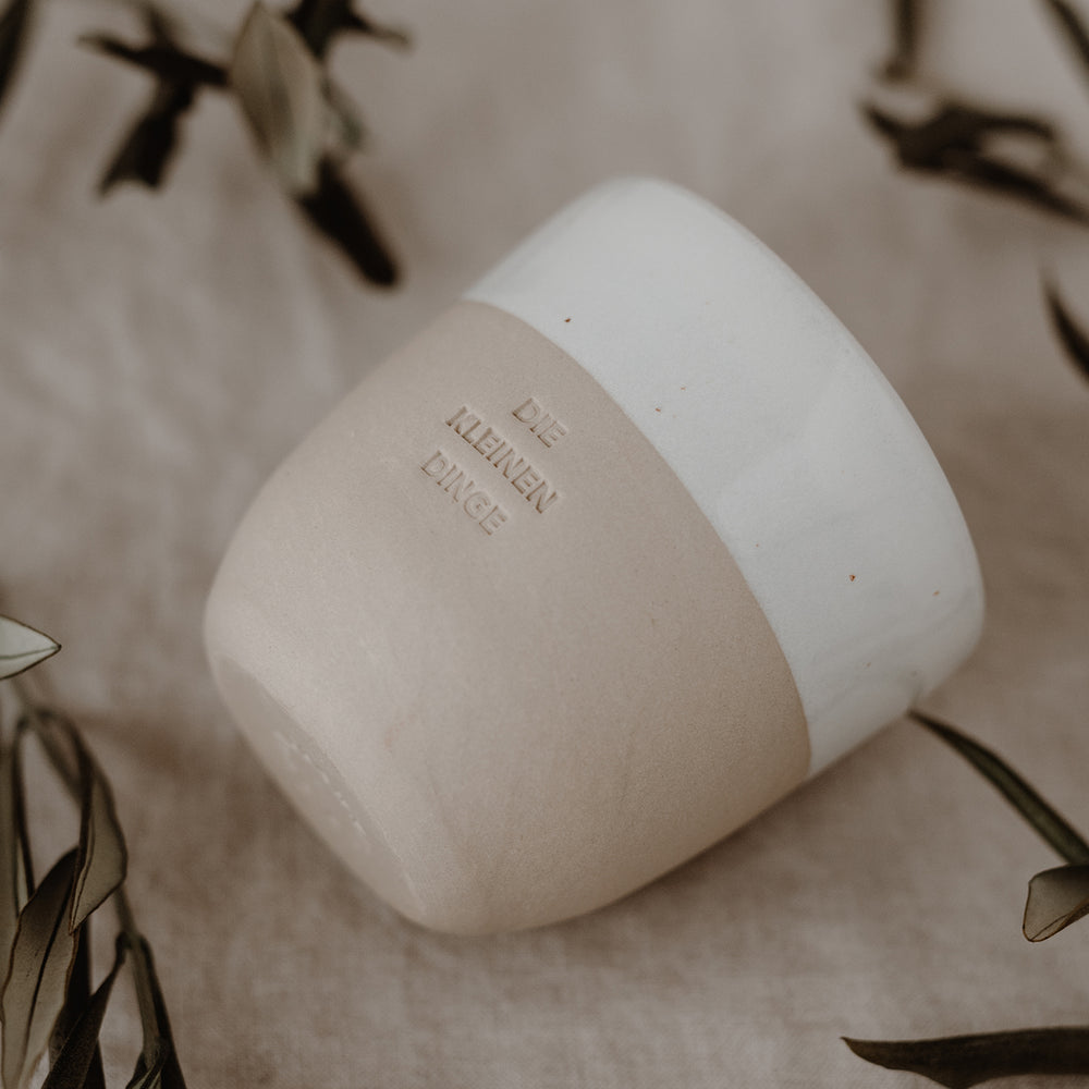 Ein heller Keramikbecher mit der Aufschrift ‚DIE KLEINEN DINGE‘ liegt auf einem Leinentuch, umgeben von grünen Zweigen.