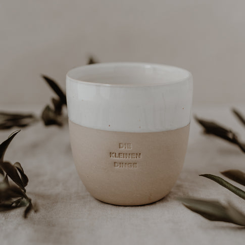 „Becher mit der Prägung ‚DIE KLEINEN DINGE‘, minimalistisch inszeniert auf hellem Untergrund mit einigen Zweigen im Bildrand.
