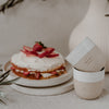 Zwei Keramikbecher, einer mit der Prägung ‚DIE KLEINEN DINGE‘, stehen neben einem Erdbeerkuchen auf einem Teller. Im Hintergrund eine Vase.