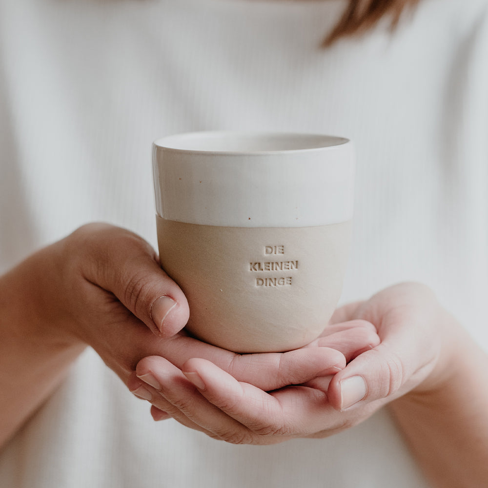 Eine Person hält einen schlichten Becher aus Keramik mit der Prägung ‚DIE KLEINEN DINGE‘ in beiden Händen. Der Hintergrund ist weich und hell.