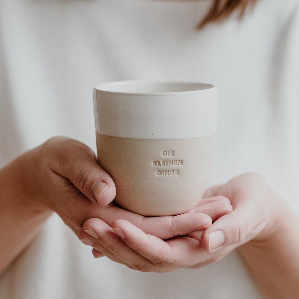Eine Person hält einen schlichten Becher aus Keramik mit der Prägung ‚DIE KLEINEN DINGE‘ in beiden Händen. Der Hintergrund ist weich und hell.