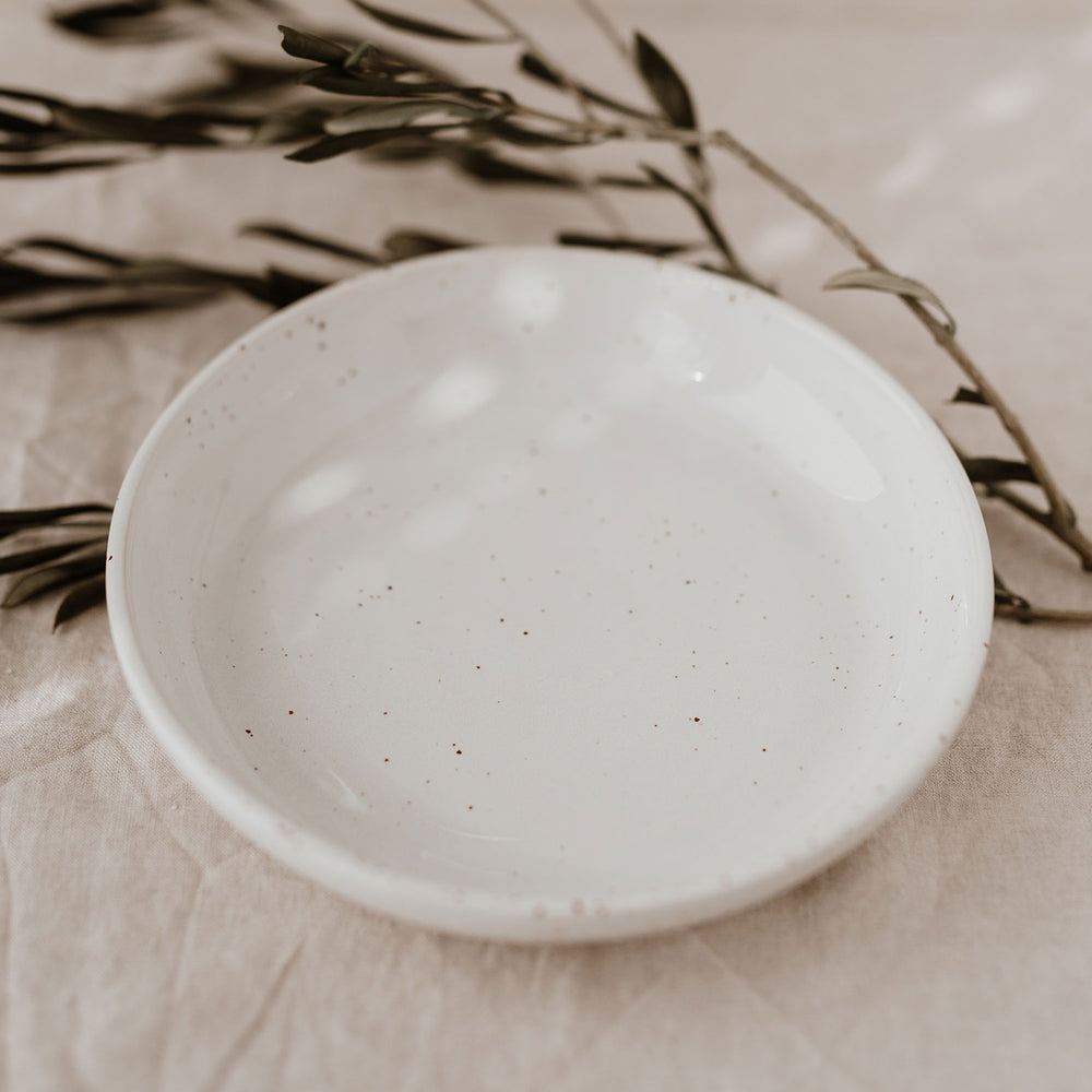 Ein einzelner tiefer Teller mit gesprenkelter Oberfläche liegt auf hellem Leinen. Olivenzweige rahmen ihn dekorativ ein.