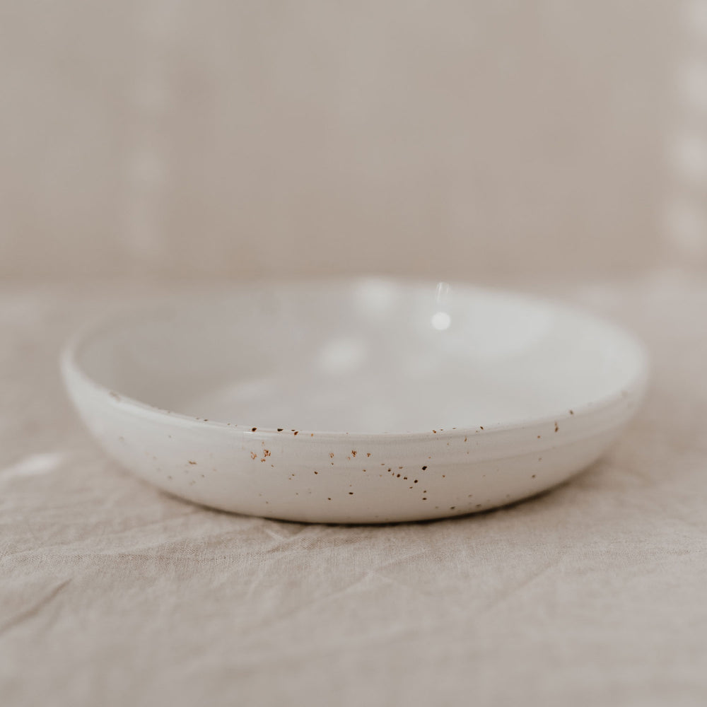 Ein tiefer Keramikteller mit gesprenkelter Glasur steht einzeln auf einem naturfarbenen Leinentuch vor hellem Hintergrund.