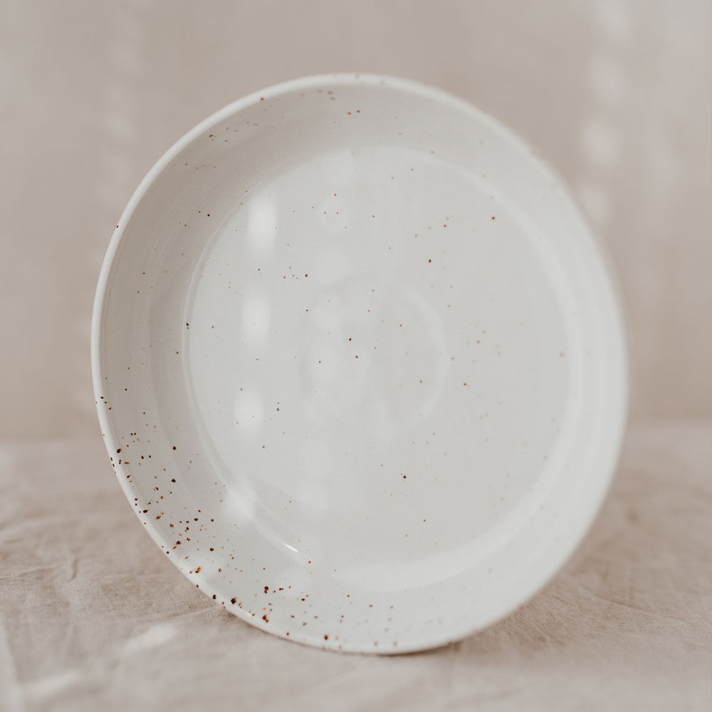 Der tiefe Teller ist frontal von oben aufgenommen und zeigt die gesprenkelte, glänzend glasierte Oberfläche auf hellem Stoff.