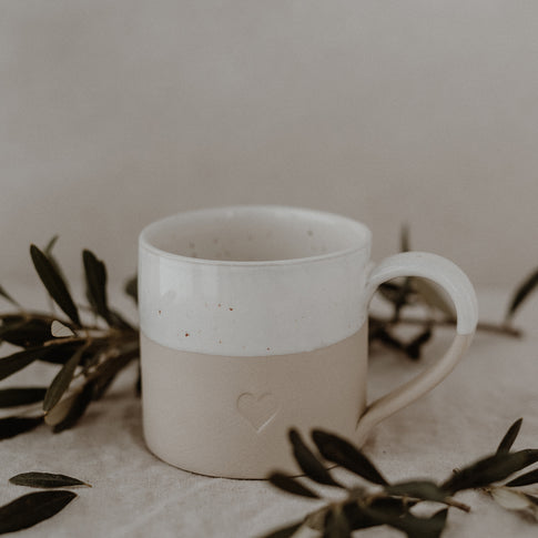 „Beige-weiße Keramiktasse mit gesprenkelter Glasur und Henkel steht auf einem Leinentuch. Auf der Vorderseite ist ein kleines, eingeprägtes Herz zu sehen. Rund um die Tasse sind Olivenzweige arrangiert – das Bild vermittelt Natürlichkeit, Achtsamkeit und handgefertigte Qualität.