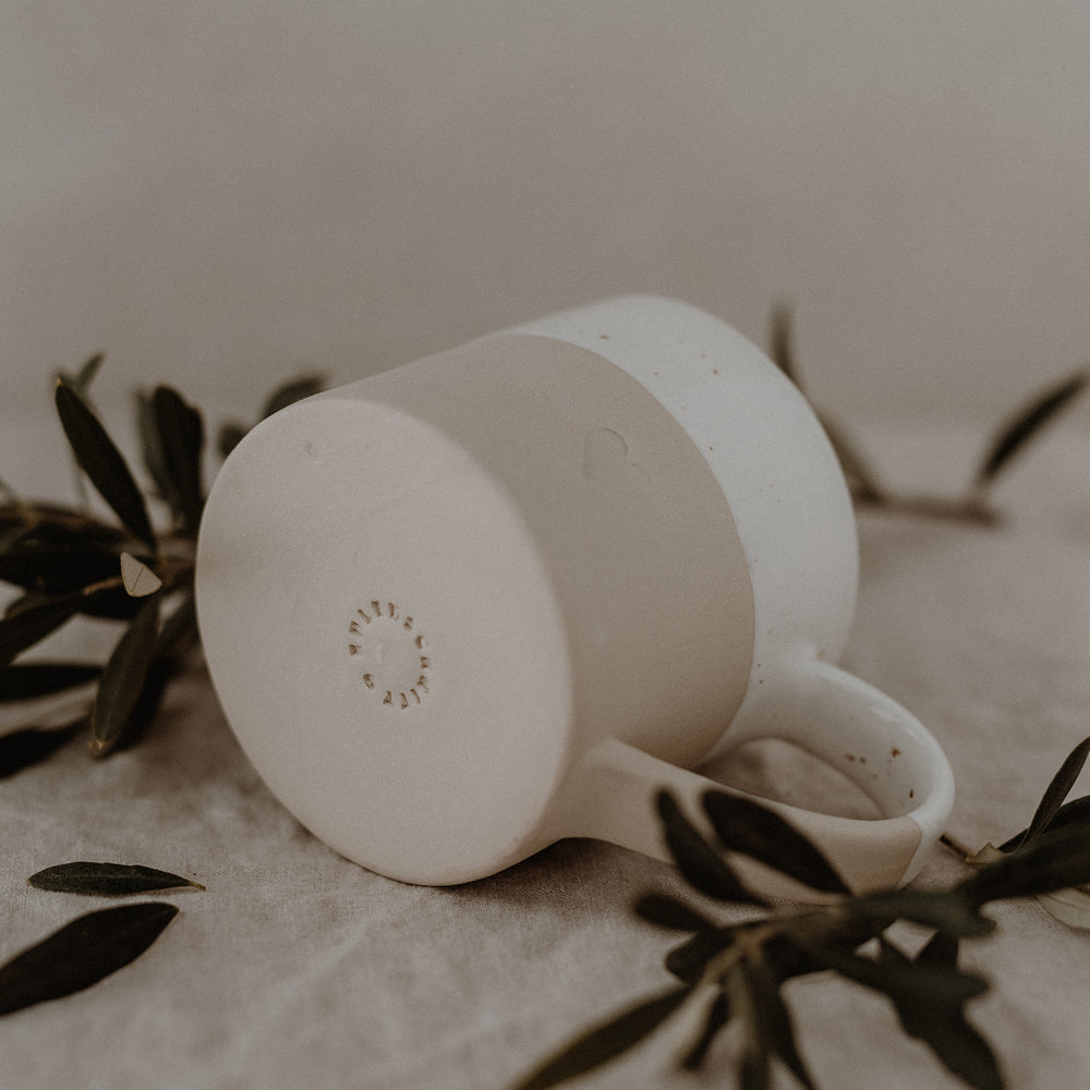 „Beige-weiße Keramiktasse liegt auf der Seite, der Boden ist sichtbar. Am Tassenboden ist der Schriftzug ‚EULENSCHNITT‘ eingraviert. Die matte untere Hälfte zeigt eine dezente Herzprägung. Olivenzweige umrahmen das Bild und verleihen eine natürliche, ruhige Stimmung.