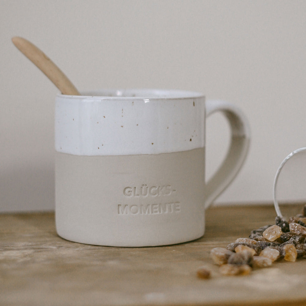 Beige-weiße Keramiktasse mit gesprenkelter Glasur, Henkel und der Gravur ‚GLÜCKSMOMENTE‘ steht auf einem rustikalen Holztisch. In der Tasse steckt ein Holzlöffel. Rechts im Bild liegen Kandisstücke – das Bild vermittelt einen gemütlichen Moment für Tee oder Kaffee.