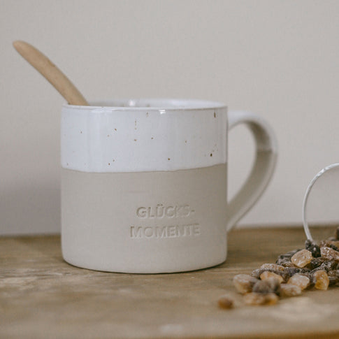 Beige-weiße Keramiktasse mit gesprenkelter Glasur, Henkel und der Gravur ‚GLÜCKSMOMENTE‘ steht auf einem rustikalen Holztisch. In der Tasse steckt ein Holzlöffel. Rechts im Bild liegen Kandisstücke – das Bild vermittelt einen gemütlichen Moment für Tee oder Kaffee.