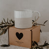 Beige-weiße Keramiktasse mit gesprenkelter Glasur und Henkel steht auf einer braunen Geschenkverpackung mit schwarzem Herzsymbol. Auf der Vorderseite der Tasse ist das Wort ‚GLÜCKSMOMENTE‘ eingeprägt. Umgeben von Olivenzweigen vermittelt die Szene eine liebevolle, natürliche Geschenkidee für besondere Augenblicke.