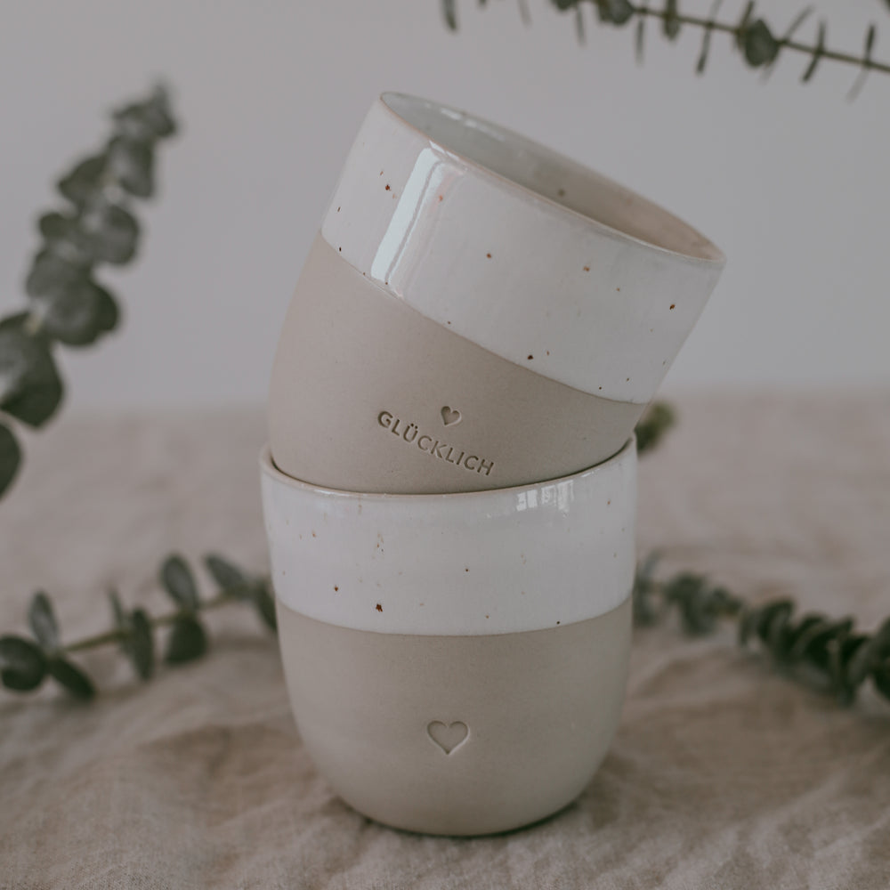 Zwei übereinandergestapelte Becher aus Keramik, verziert mit Herzsymbol und dem Wort ‚GLÜCKLICH‘. Im Hintergrund zarte Eukalyptuszweige.