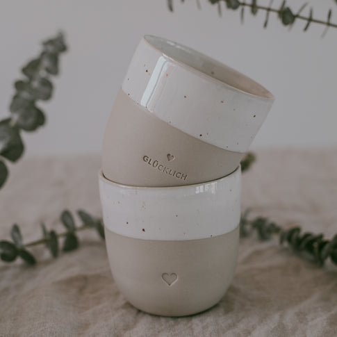 Zwei übereinandergestapelte Becher aus Keramik, verziert mit Herzsymbol und dem Wort ‚GLÜCKLICH‘. Im Hintergrund zarte Eukalyptuszweige.