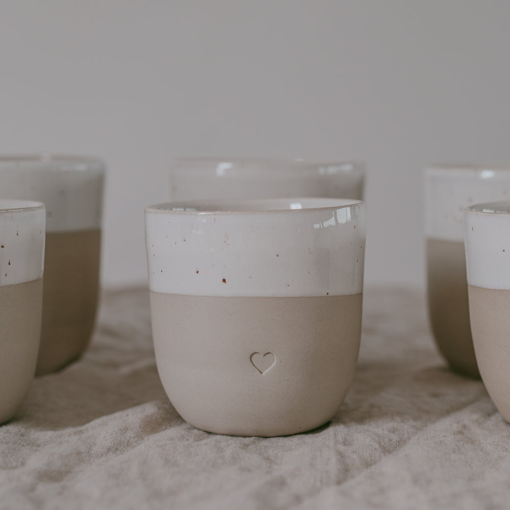 Becher aus matter Keramik mit glänzend glasierter Oberkante und geprägtem Herz. Mehrere Becher stehen locker arrangiert auf einem strukturierten Tuch.