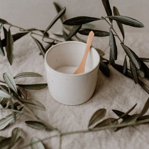 Kleiner, heller Keramikbecher mit Holzlöffel, eingefasst von Olivenzweigen auf einem naturfarbenen Leinentuch. Die Oberfläche ist fein gesprenkelt.