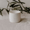 Schlichter, zylindrischer Keramikbecher mit feiner brauner Sprenkelung auf hellem Grund. Im Hintergrund sind Olivenzweige stilvoll arrangiert.