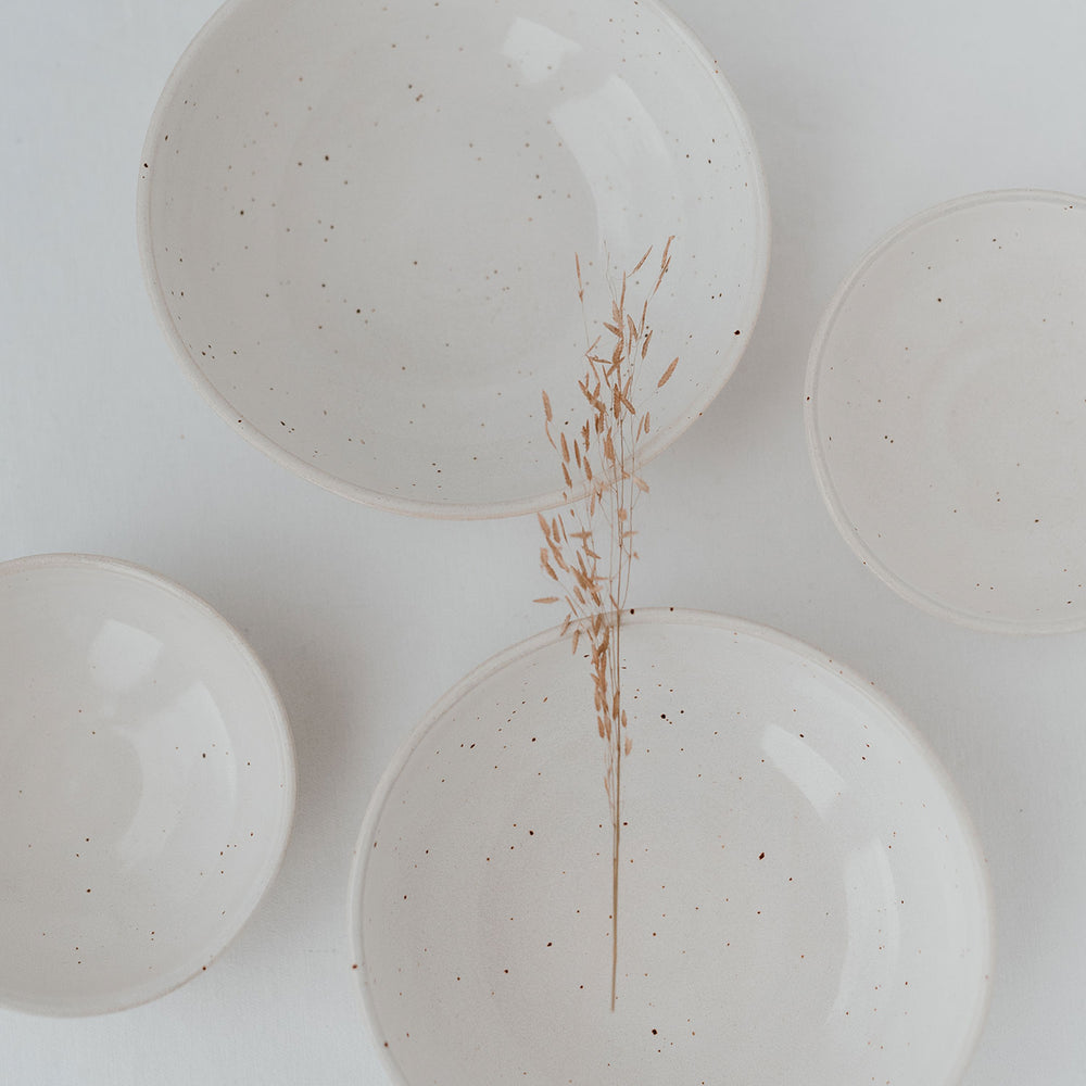 Fünf handgefertigte Keramikschalen in verschiedenen Größen mit cremefarbener Glasur und feinen Sprenkeln, auf hellem Untergrund arrangiert; in der Mitte liegt ein filigraner, getrockneter Grashalm.