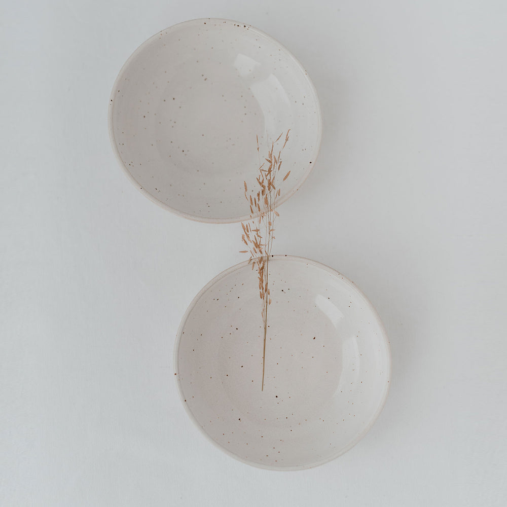 Zwei minimalistische Keramikschalen in Naturweiß mit zarten Sprenkeln, auf hellem Untergrund arrangiert, getrennt durch einen einzelnen getrockneten Grashalm – harmonisch und ruhig inszeniert.
