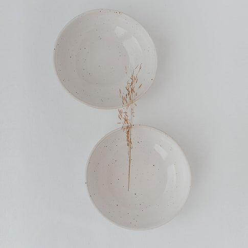 Zwei minimalistische Keramikschalen in Naturweiß mit zarten Sprenkeln, auf hellem Untergrund arrangiert, getrennt durch einen einzelnen getrockneten Grashalm – harmonisch und ruhig inszeniert.