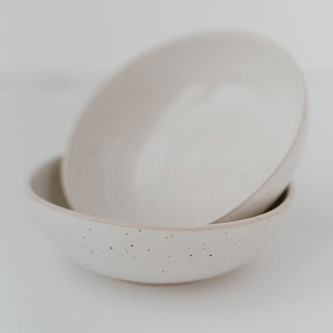 Zwei minimalistische Keramikschalen in Naturweiß mit feinen, unregelmäßigen Sprenkeln, übereinandergestapelt auf hellem Untergrund im Studio-Stil.