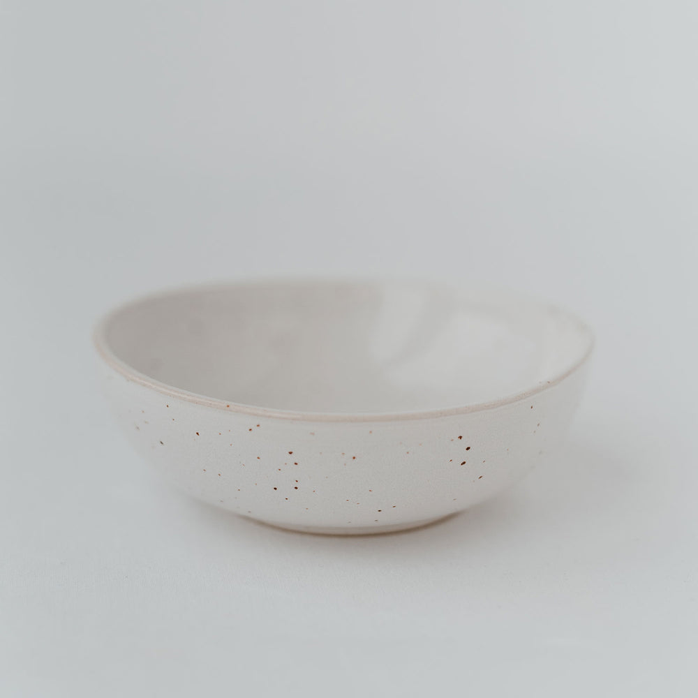 Eine einzelne handgefertigte Keramikschale in hellem Cremeweiß mit feiner, gesprenkelter Glasur steht auf einem weißen Untergrund, minimalistisch und elegant inszeniert.