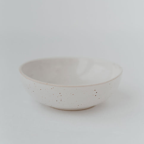 Eine einzelne handgefertigte Keramikschale in hellem Cremeweiß mit feiner, gesprenkelter Glasur steht auf einem weißen Untergrund, minimalistisch und elegant inszeniert.