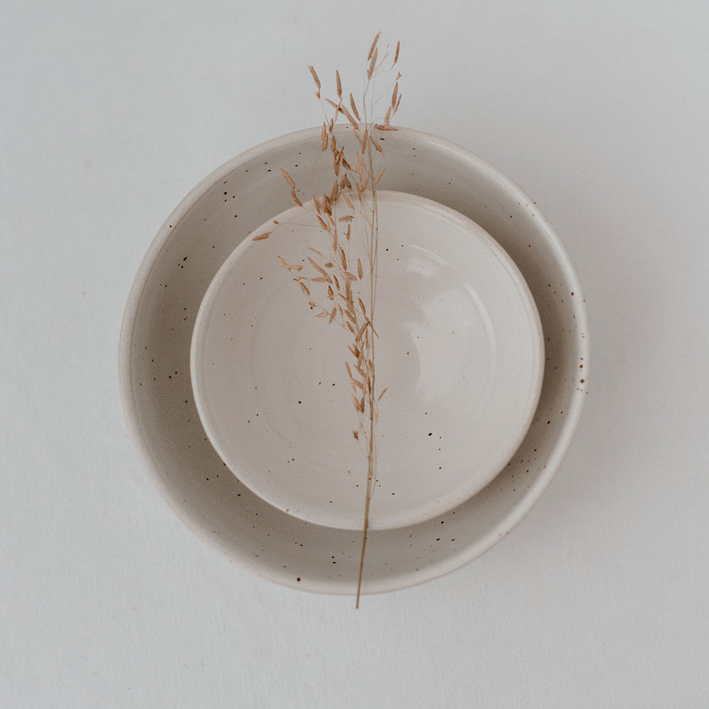 Zwei handgefertigte, cremefarbene Keramikschalen mit feiner Sprenkelung sind ineinander gestapelt; darüber liegt ein filigraner, getrockneter Grashalm auf weißem Hintergrund – minimalistisch und ästhetisch arrangiert.