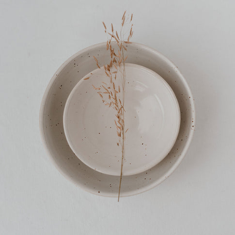 Zwei handgefertigte, cremefarbene Keramikschalen mit feiner Sprenkelung sind ineinander gestapelt; darüber liegt ein filigraner, getrockneter Grashalm auf weißem Hintergrund – minimalistisch und ästhetisch arrangiert.