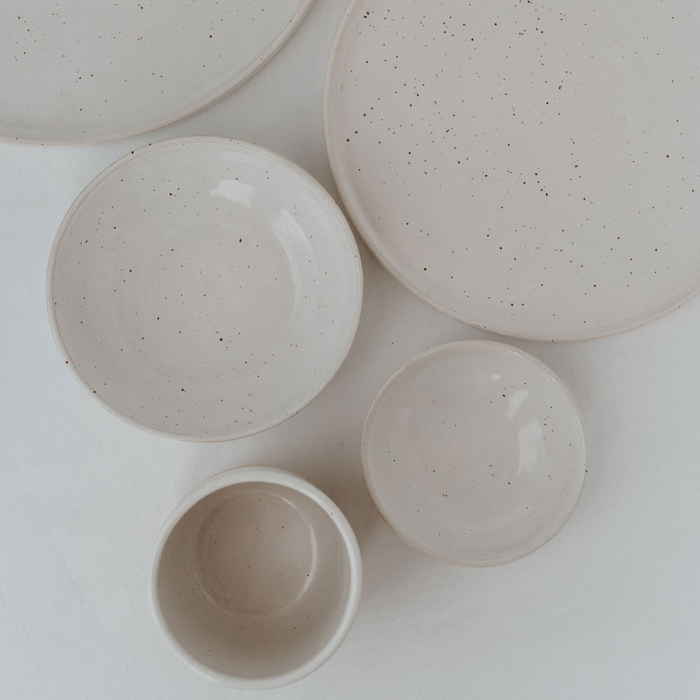 Eine flache Anordnung aus handgefertigten, gesprenkelten Keramikschalen und -tellern in verschiedenen Größen auf weißem Leinentuch – minimalistisches, natürliches Geschirrdesign in heller Farbgebung.