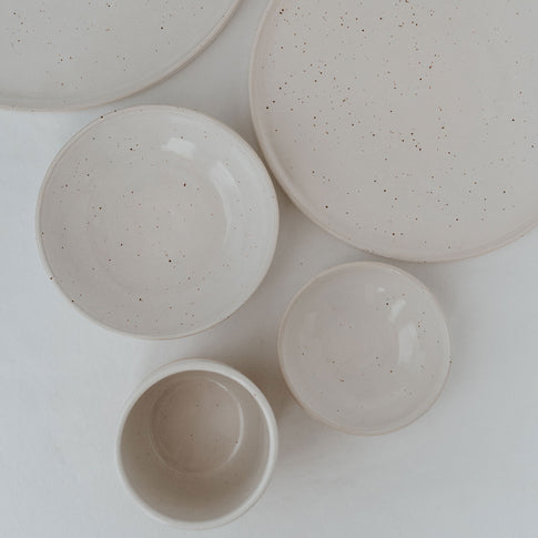 Eine flache Anordnung aus handgefertigten, gesprenkelten Keramikschalen und -tellern in verschiedenen Größen auf weißem Leinentuch – minimalistisches, natürliches Geschirrdesign in heller Farbgebung.