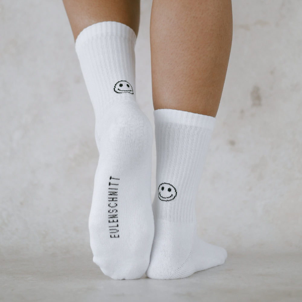 Person trägt weiße Socken mit schwarzem Smiley-Motiv und „Eulenschnitt“-Schriftzug auf der Sohle, vor hellem Hintergrund. Nahaufnahme der Socken und Beine.