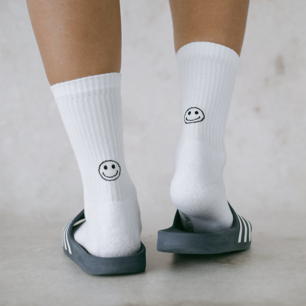 Nahaufnahme von zwei Beinen in weißen Socken mit schwarzem Smiley-Motiv auf der Rückseite, getragen in dunkleblauen Adiletten, stehend auf hellem Boden.